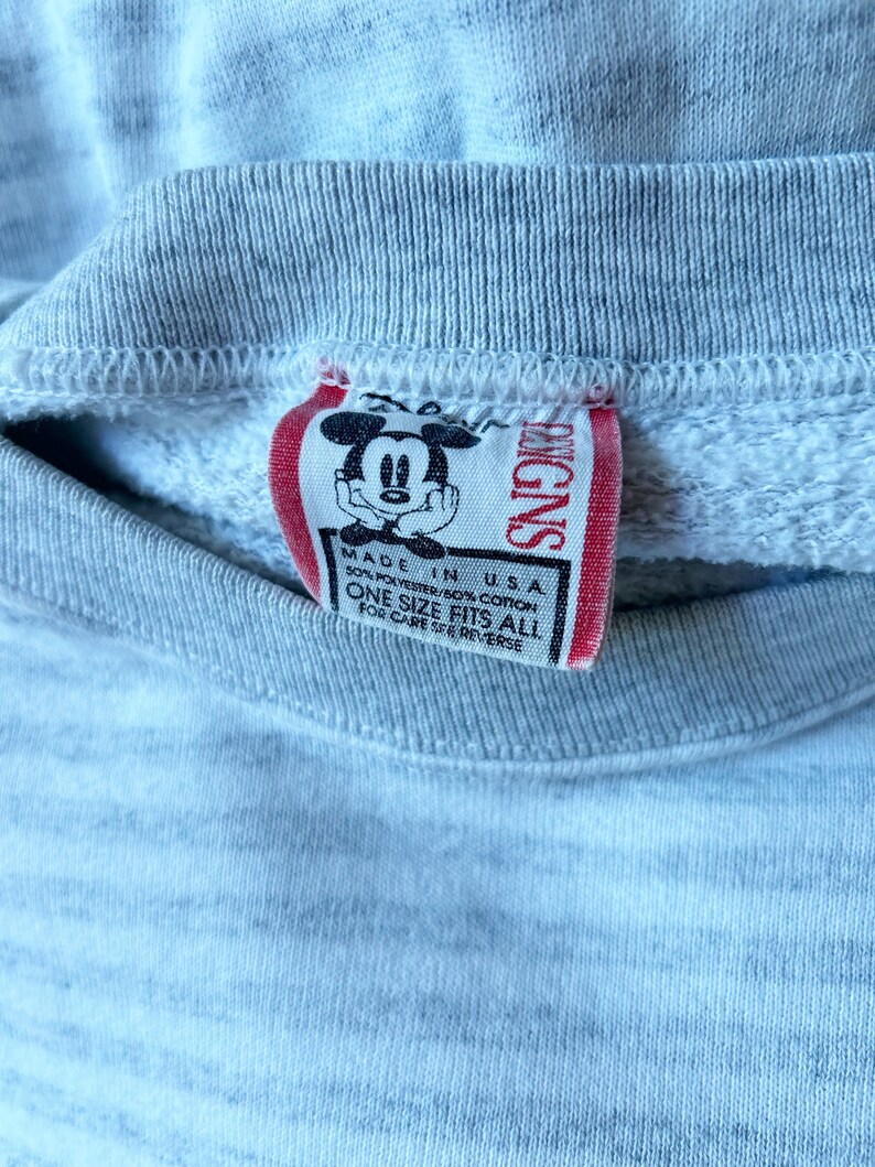 May include: A gray sweatshirt with a red tag featuring a cartoon Mickey Mouse. The tag reads "Disney" and "Made in USA 50% Polyester/50% Cotton One Size Fits All For Care See Reverse."