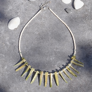 May include: A delicate necklace featuring a white beaded strand with a series of light green, elongated shell-like pendants. The necklace is displayed on a gray surface, accompanied by seashells and small stones.