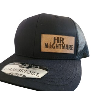 Op de afbeelding: Zwarte truckerpet met een mesh achterkant en een gebogen klep. Een rechthoekige bruine leren patch toont de tekst "HR NIGHTMARE" met een middelvinger-afbeelding. De pet heeft een "CAMBRIDGE" label.