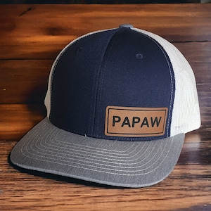 May include: A navy blue and gray baseball cap with a white mesh back. The cap features a gray brim and a leather patch with the word "PAPAW" in capital letters.