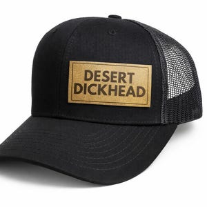 May include: Black trucker hat with a mesh back and a curved brim. The hat features a rectangular patch with the words "DESERT DICKHEAD" in a brown, bold font. The hat is designed for casual wear.