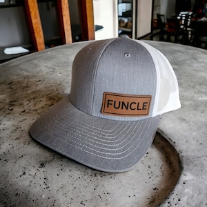 May include: A gray baseball cap with a white mesh back. The cap features a brown leather patch with the word "FUncle" in black block letters. The hat is sitting on a gray surface.