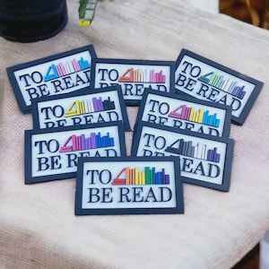 May include: A collection of black framed magnets, each displaying the text "TO BE READ" in white. The magnets feature a colourful stack of books. The books are in various colours, including pink, blue, yellow, and purple.