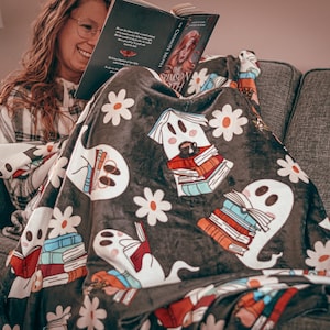 Peut inclure: Une couverture gris foncé avec un motif de fantômes blancs, de livres et de marguerites. La couverture est drapée sur une personne assise sur un canapé gris, lisant un livre. La couverture du livre est visible.