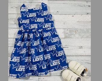May include: A blue sleeveless dress with a white print of the Detroit Lions logo repeated all over. The dress has a gathered skirt. Two white shoes are on the wooden floor.