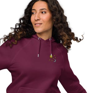 May include: A maroon hoodie with a drawstring hood and a kangaroo pocket. The hoodie features an embroidered design of a sun, stars, and a crescent moon on the upper left chest. The hoodie is made of a soft material.