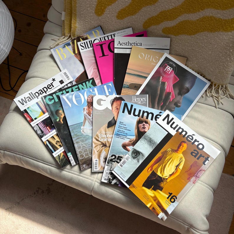 May include: A collection of fashion and art magazines, including Vogue, Numero, and Wallpaper, are fanned out on a white cushioned surface. The magazines feature colorful covers with various models and graphic designs. The magazines are arranged in a casual, overlapping manner.