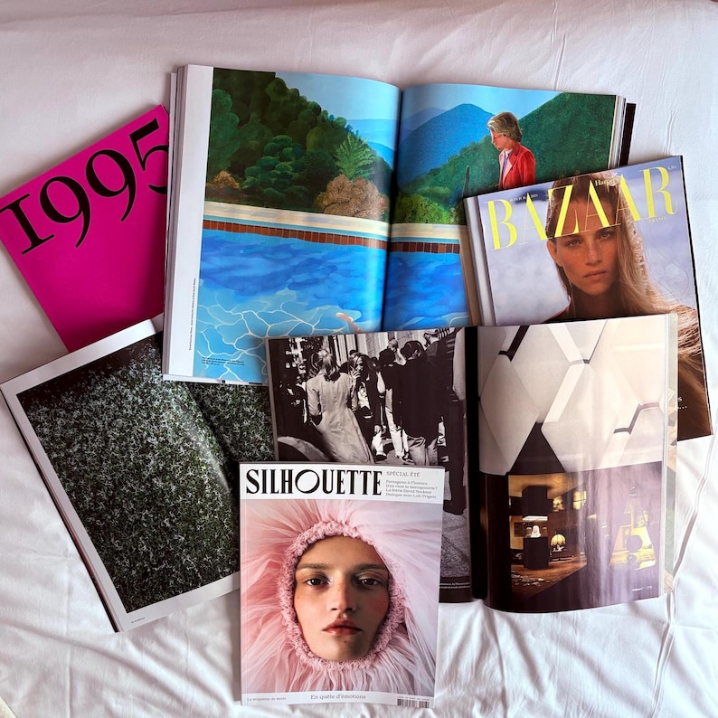 May include: A collection of open magazines and fashion publications, including a pink cover with "1995" in large black font. Other magazines include "BAZAAR" and "SILHOUETTE", with images of models and art.