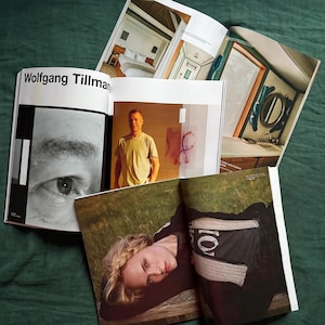 May include: Several open magazines are displayed on a green surface. One magazine features the text "Wolfgang Tillmans" and a black and white close-up of an eye. Other magazines show interior design and a portrait of a person.