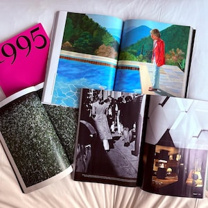 May include: An assortment of open magazines and a pink book with the number "995" printed in black. The magazines display colorful and black and white photographs, including a pool scene and a crowd of people.