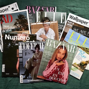 May include: A collection of fashion and design magazines, including "Wallpaper," "AD," "Bazaar," and "Elle Decoration," are arranged on a dark green surface. The covers feature various models, text, and imagery, creating a visually rich display.