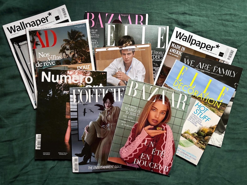 May include: A collection of fashion and design magazines, including "Wallpaper," "AD," "Bazaar," and "Elle Decoration," are arranged on a dark green surface. The covers feature various models, text, and imagery, creating a visually rich display.