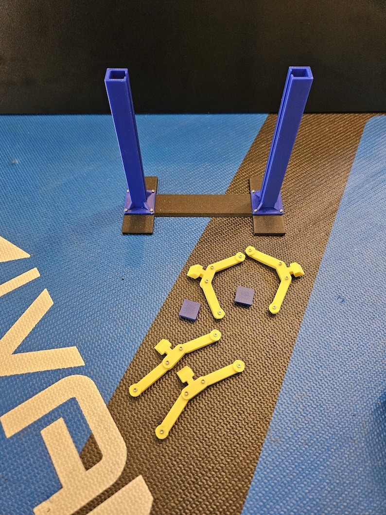 May include: A disassembled, 3D-printed structure with blue vertical supports and yellow, curved components. The base is black, and the components are arranged on a blue and black patterned surface. Small purple squares are also present.