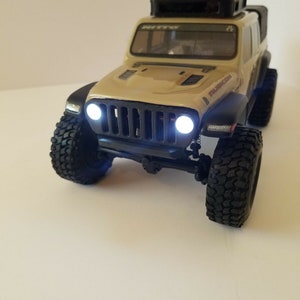 May include: A tan Jeep Wrangler Rubicon toy truck with black wheels and a black roof rack. The truck has headlights that are turned on and a black bumper.