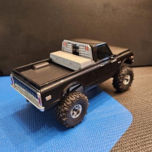 May include: A black toy pickup truck with large off-road tires. The truck has a gray toolbox and a silver front bumper. The truck is on a blue surface.