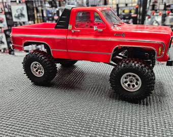 May include: A red toy pickup truck with a black cab and a silver trim. The truck has black wheels with silver rims and is sitting on a gray textured surface.