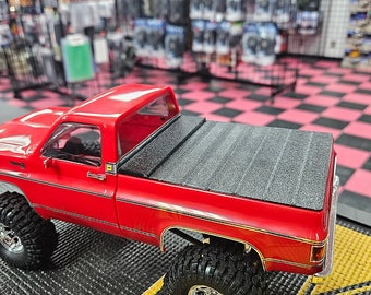May include: Red toy pickup truck with a black bed cover. The truck has large black tires and a chrome bumper. The truck is sitting on a black and yellow checkered mat.