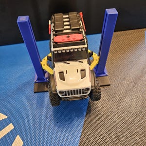 May include: A white toy Jeep with a black roof rack and red accents is raised on a blue and yellow toy car lift. The Jeep has black tires and a white front grill. The lift has two blue posts.