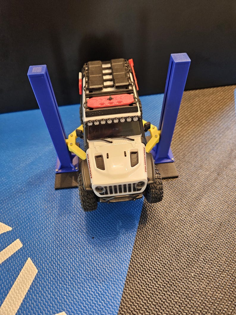 May include: A white toy Jeep with a black roof rack and red accents is raised on a blue and yellow toy car lift. The Jeep has black tires and a white front grill. The lift has two blue posts.