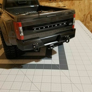 May include: A black model pickup truck with the word "PLATINUM" in white lettering across the tailgate. The truck has a black bumper with two tow hooks and a trailer hitch. The truck is on a gray and white grid surface.