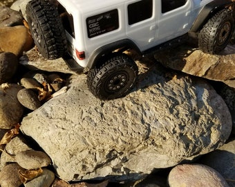 May include: White Jeep Wrangler Rubicon toy car with black tires and KMC wheels. The car is on a pile of rocks.