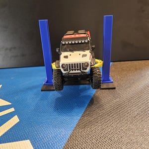May include: A white and black toy Jeep Wrangler on a blue and yellow car lift. The Jeep has a black grill, black tires, and a red roof rack. The lift has two blue support columns and a yellow base.