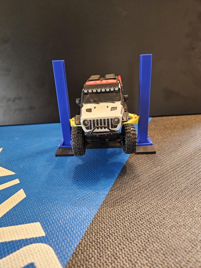 May include: A white and black toy Jeep Wrangler on a blue and yellow car lift. The Jeep has a black grill, black tires, and a red roof rack. The lift has two blue support columns and a yellow base.