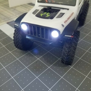 May include: White Jeep Rubicon toy car with black accents and headlights on. The car is sitting on a gray grid surface.