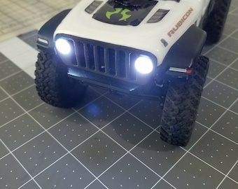 May include: White Jeep Rubicon toy car with black accents and headlights on. The car is sitting on a gray grid surface.