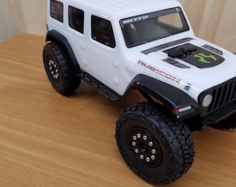 May include: White Jeep Wrangler Rubicon RC car with black wheels and tires. The car has a black hood with a green and black graphic. The car has a black windshield with the word "NITTO" printed on it.