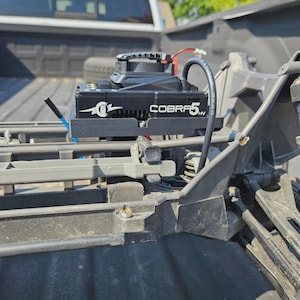 May include: Black "COBRA5" electronic speed controller for a remote control vehicle. The controller is mounted on a gray chassis and has a fan and wiring. The image is taken outdoors in daylight.