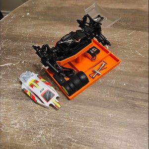 May include: An orange and black plastic storage tray with a black RC car chassis, four black tires, and tools. The tray is on a wooden surface.