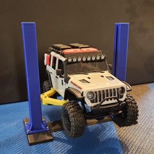 May include: A white and black toy Jeep Rubicon with black off-road tires is raised on a yellow and blue toy car lift. The Jeep has a roof rack and a light bar. The word "RUBICON" is visible on the hood.
