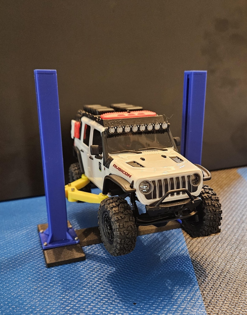 May include: A white and black toy Jeep Rubicon with black off-road tires is raised on a yellow and blue toy car lift. The Jeep has a roof rack and a light bar. The word "RUBICON" is visible on the hood.