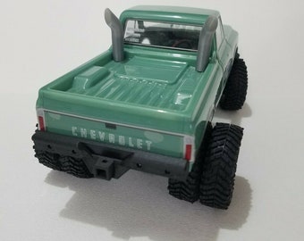 May include: A green Chevrolet pickup truck model with a black rear bumper and large tires. The truck has two exhaust pipes that extend up from the bed.