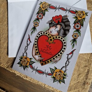 May include: Valentine's Day card with a vintage tattoo design. The card features a woman holding a red heart with the words "My Valentine." The design includes yellow roses, barbed wire, and a heart pierced by an arrow. The card is inside a white envelope.