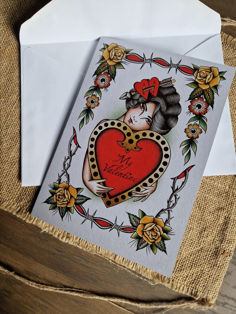 May include: Valentine's Day card with a vintage tattoo design. The card features a woman holding a red heart with the words "My Valentine." The design includes yellow roses, barbed wire, and a heart pierced by an arrow. The card is inside a white envelope.