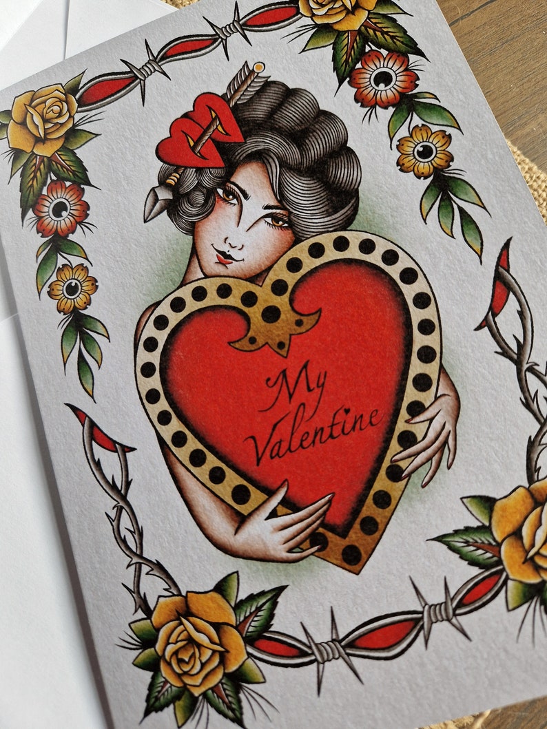 May include: Greeting card featuring a vintage tattoo-style illustration. A woman with dark hair and a red heart-shaped hair accessory embraces a large red heart with the words "My Valentine." The design includes yellow roses, barbed wire, and a decorative border.