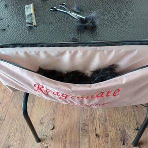Op de afbeelding: Een opstelling voor het verzorgen van huisdieren met een zwarte tafel. Een roze stoffen zak met het rode woord "Reagrenate" is bevestigd. Schaar, kam en zwart haar liggen op de tafel, wat wijst op recente verzorging.