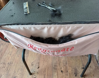 May include: A pet grooming setup featuring a black table with a black surface. A pink fabric bag with the word "Reagrenate" in red is attached to the table. Scissors, a comb, and black fur are on the table.