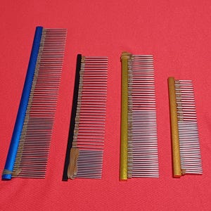 Könnte beinhalten: Vier Pflegekämme mit metallisch silbernen Zinken und farbigen Griffen. Die Griffe sind blau, schwarz, gold und ein kürzerer goldener Griff. Jeder Kamm hat kleine Gummibänder, die um die Basis der Zinken gewickelt sind, um sie am Griff zu befestigen.