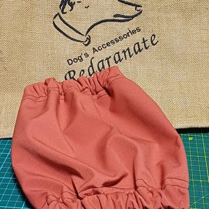 May include: A rust-colored dog accessory, possibly a snood or bandana, with a gathered elastic edge. The background features a burlap bag with a dog head logo and the text "Dog's Accessories Redaranate". The accessory is on a green cutting mat.