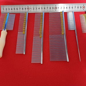May include: Set of seven dog grooming combs, including a comb with a wooden handle. The combs are made of metal and have different sized teeth. The combs are laid out on a red surface with a ruler for scale.