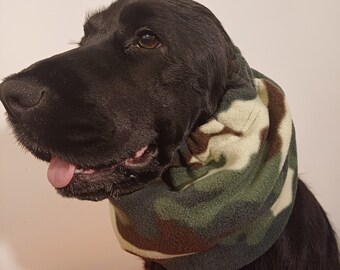 May include: Black dog wearing a camouflage fleece neck warmer. The dog is looking to the left and has its tongue sticking out.