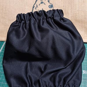 以下が含まれることがあります： 上部と下部に伸縮性のあるギャザー開口部を備えた、質感のある生地で作られた黒い犬用スヌード。スヌードは、Redgranate snoods for dogsという刺繍テキストが施されたベージュの麻生地の前に配置されています。