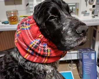 May include: A black and white dog wearing a red plaid fleece scarf. The dog has a long snout and is looking to the side. The background includes a sewing machine and other sewing supplies.