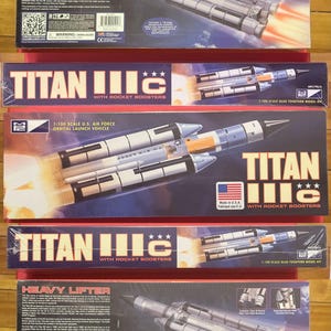 May include: Multiple model kits of the Titan III C rocket, with rocket boosters, and the Heavy Lifter. The boxes feature illustrations of the rockets in flight, with flames and smoke. The boxes are red, blue, and black.