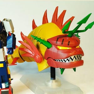 以下が含まれることがあります： ロボットとドラゴンに似た生き物のカラフルなアクションフィギュア。ロボットは主に青、白、赤で、黄色のアクセントがあります。ドラゴンは黄色、赤、緑で、鋭い歯ととげのあるデザインです。両方のフィギュアは透明なスタンドにあります。