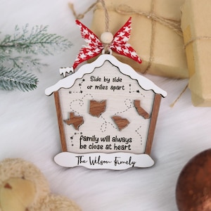 May include: A wooden house-shaped ornament with a white roof and brown frame. The ornament features the text "Side by side or miles apart" and "family will always be close at heart." It includes state outlines and is topped with a red and white bow.
