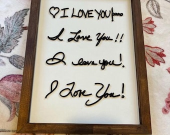 May include: A framed wooden sign with a white background. The sign features the phrase "I Love You" written in black cursive script four times. The frame is a dark brown color.